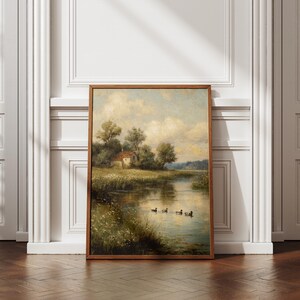 May include: A framed landscape painting depicts a serene waterside scene. A small house sits among trees on the bank, with ducks swimming in the water. The artwork is in a wooden frame, leaning against a white wall and a dark wood floor.
