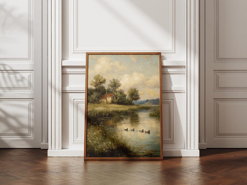 May include: A framed landscape painting depicts a serene waterside scene. A small house sits among trees on the bank, with ducks swimming in the water. The artwork is in a wooden frame, leaning against a white wall and a dark wood floor.