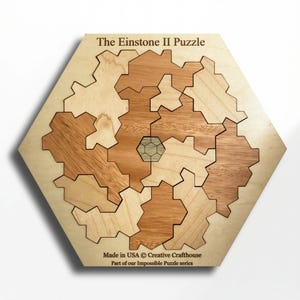 May include: The Einstone II Puzzle, a wooden jigsaw puzzle with a unique gear-like design. The puzzle is made of light and dark wood pieces, with a hexagonal frame. The text "The Einstone II Puzzle" is at the top. Made in the USA.