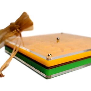 May include: A square, multi-layered acrylic box with a clear top, yellow, brown, and green layers. A small gold-colored drawstring bag is to the left. The box has gold-colored corner accents and a small silver ball on top.