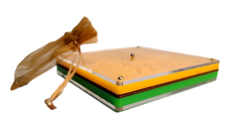 May include: A square, multi-layered acrylic box with a clear top, yellow, brown, and green layers. A small gold-colored drawstring bag is to the left. The box has gold-colored corner accents and a small silver ball on top.