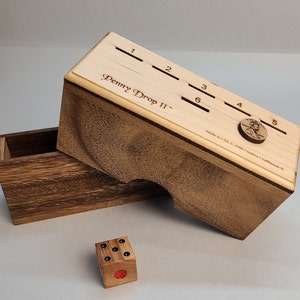 Pode incluir: Jogo Penny Drop II de madeira com uma caixa de madeira e um dado de seis lados. O jogo tem ranhuras numeradas de 1 a 6. A caixa é feita nos EUA pela Creative Crafthouse.