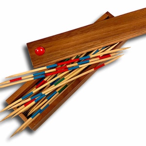 May include: Wooden pick-up sticks game set in a rectangular wooden box. The sticks are painted with red, blue, and green bands. The box has a red circular accent on the lid. The game is open, with the sticks partially spilling out.