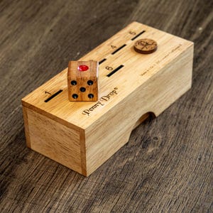 May include: Wooden Penny Drop game with a dice and a wooden coin. The game box is light brown with the numbers 1 through 6 etched on the top. The dice is brown with black dots and a red dot. The game is on a wooden surface.