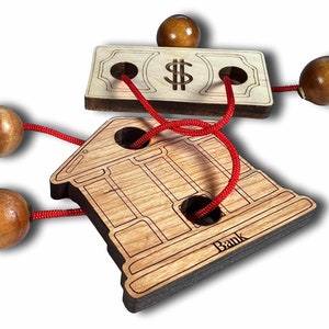 May include: Wooden puzzle with a bank building, a dollar bill, and four wooden balls connected by a red string. The bank building has the word "Bank" inscribed on the bottom.