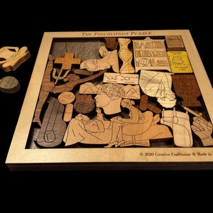May include: The Psychiatrist Puzzle is a wooden jigsaw puzzle with a variety of shapes and designs. The puzzle features a psychiatrist and patient, as well as other symbols related to mental health. The puzzle is made in the USA. &copy; 2020 Creative Crafthouse.