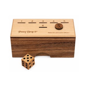 May include: A rectangular wooden game box labeled Penny Drop II with slots numbered 1 through 6 on the top surface. A single copper penny rests near slot 4. A wooden six-sided die with black pips sits in front of the box. The text Made in USA 2018 Creative Crafthouse is printed on the top.