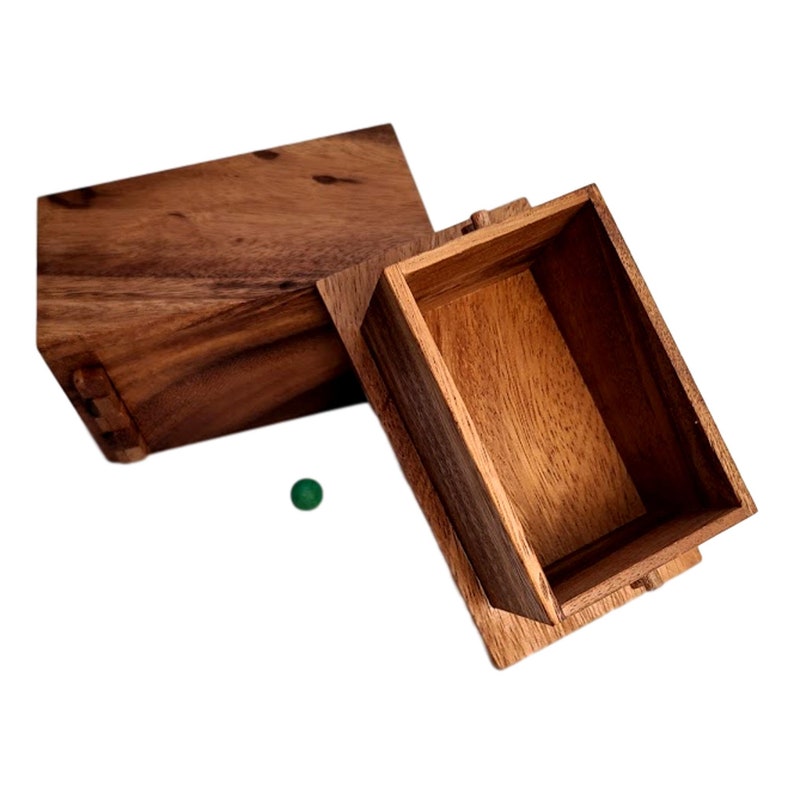 May include: A wooden box with a hinged lid, open to reveal an empty interior. The box is made of dark brown wood with a natural grain pattern. A small green ball is visible in the foreground.