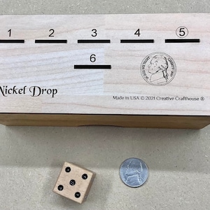 May include: A wooden box with the words "Nickel Drop" engraved on the front. The box has six slots numbered 1 through 6. A nickel is visible in the slot labeled 5. The box is made in the USA by Creative Crafthouse. A wooden dice with black dots is on a light brown surface next to a silver coin.