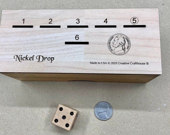 May include: A wooden box with the words "Nickel Drop" engraved on the front. The box has six slots numbered 1 through 6. A nickel is visible in the slot labeled 5. The box is made in the USA by Creative Crafthouse. A wooden dice with black dots is on a light brown surface next to a silver coin.