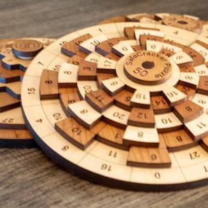Puede incluir: Un rompecabezas de caja fuerte de madera con un diseño circular. El rompecabezas tiene múltiples capas de anillos concéntricos con números en cada anillo. El centro del rompecabezas tiene una pieza circular con el texto "Safe Cracker Puzzle" y el número "50".