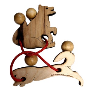 May include: Wooden puzzle featuring a wolf and a rabbit, both with red string and wooden beads threaded through them. The string is tied in a knot and the animals are positioned so that the wolf is on top of the rabbit. The puzzle is made in the USA by Creative Crafthouse.