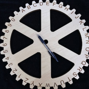 Może przedstawiać: Zębatka drewniana z numerami od 0 do 9 i literami od A do Z wokół krawędzi. Zębatka ma sześć szprych i leży na niej czarny długopis.
