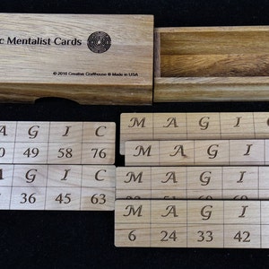 May include: Wooden box with a set of five wooden tiles. Each tile has the letters M, A, G, I, and C printed on it. The tiles also have numbers printed on them, such as 22, 40, 49, 58, and 76. The box has the words "Magic Mentalist Cards" printed on it.