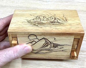 Op de afbeelding: Een houten puzzeldoos met een schuifmechanisme. De doos is versierd met een gegraveerd ontwerp van bergen en een zon. De doos is gemaakt van licht hout met een natuurlijke afwerking.