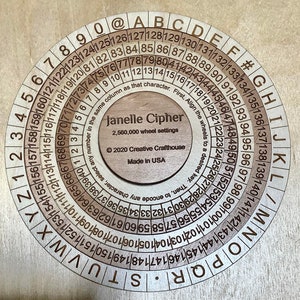 Puede incluir: Una rueda de cifrado circular de madera con el texto "Janelle Cipher" y "2.560.000 ajustes de rueda" en el centro. La rueda tiene una serie de círculos concéntricos con números y letras alrededor del borde. El texto "© 2020 Creative Crafthouse Made in USA" también es visible.