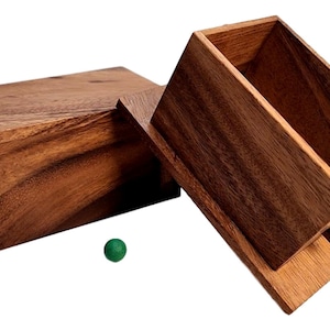 May include: A wooden box with a hinged lid, open to reveal the interior. The box is made of dark brown wood with a natural grain pattern. A small green ball sits on the table next to the box.