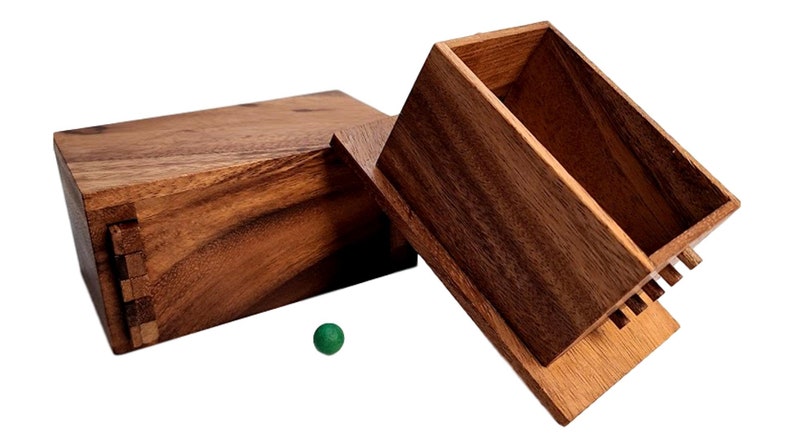 May include: A wooden box with a hinged lid, open to reveal the interior. The box is made of dark brown wood with a natural grain pattern. A small green ball sits on the table next to the box.