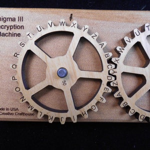 May include: Wooden Enigma III Encryption Machine featuring two gear wheels, each inscribed with the alphabet. The text "Made in USA" and "Creative Crafthouse" are present. The gears are mounted on a light brown wooden base.