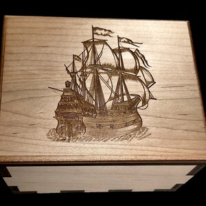 Könnte beinhalten: Eine Holzkiste mit einem lasergravierten Bild eines Segelschiffs mit mehreren Segeln. Das Schiff befindet sich in der Mitte des Kistendeckels und ist von einem hellen Holzhintergrund umgeben.