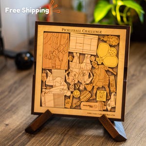 May include: A wooden pickleball challenge puzzle with a brown frame and stand. The puzzle features a variety of pickleball-themed images, including players, paddles, and a net. The puzzle is made in the USA.