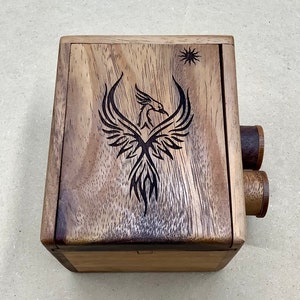 May include: A wooden box with a carved phoenix design and a sun symbol. The box is made of light and dark brown wood and has two cylindrical knobs on the side. The phoenix is black and has detailed feather patterns.