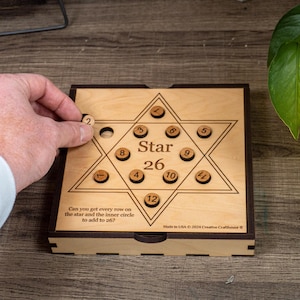 Könnte beinhalten: Ein sternförmiges Holzpuzzle-Spiel mit den Worten "Star 26" darauf gedruckt. Das Spiel enthält 12 nummerierte Holzscheiben, die auf dem Stern bewegt werden können, um zu versuchen, jede Reihe auf 26 zu addieren. Das Spiel wird in den USA von Creative Crafthouse hergestellt.