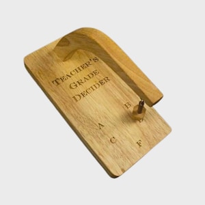 May include: A wooden "Teacher's Grade Decider" with a curved handle. The rectangular base has the text "TEACHER'S GRADE DECIDER" engraved, along with the letters A, B, C, and F. A small, wooden spinner is attached.