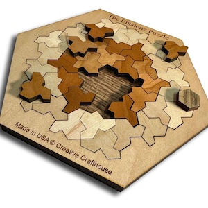 Puede incluir: Rompecabezas de madera titulado "The Einstone Puzzle" con formas geométricas entrelazadas en tonos marrones y beige. El rompecabezas está hecho en los EE. UU. por Creative Crafthouse. El rompecabezas está sobre una base de madera marrón claro.