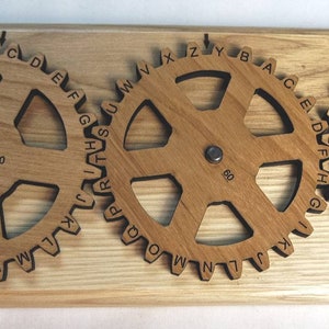 Puede incluir: Una máquina de encriptación Enigma II de madera con tres engranajes rotativos. Cada engranaje tiene un alfabeto diferente impreso en él. Los engranajes están etiquetados con los números 70, 60 y 19. La máquina está hecha en los Estados Unidos por Creative Crafthouse.