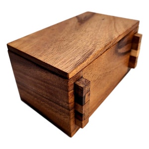 May include: A wooden box with a sliding puzzle lid. The box is made of dark brown wood with a natural grain pattern. The lid is made of several pieces of wood that slide together to open the box.