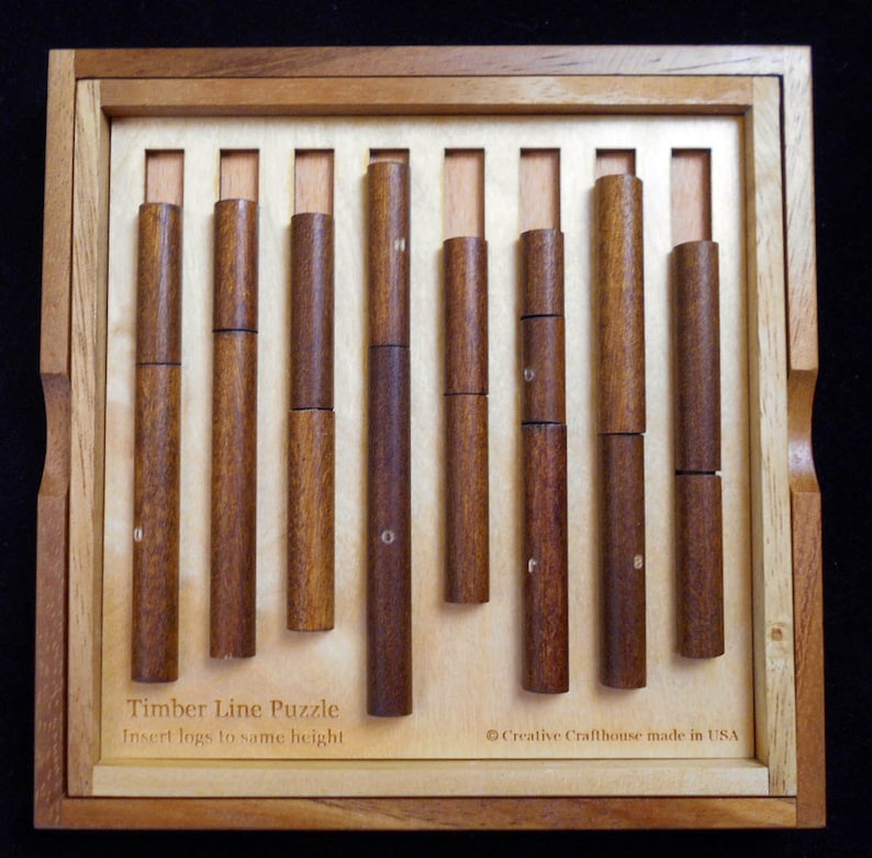 Pode incluir: Uma caixa de madeira com um conjunto de pe&ccedil;as de quebra-cabe&ccedil;a de madeira. A caixa &eacute; etiquetada como "Timber Line Puzzle" e "Insertar troncos na mesma altura". A caixa &eacute; feita nos Estados Unidos pela Creative Crafthouse.