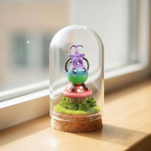 Puede incluir: Una escena en miniatura caprichosa con una criatura colorida con cabeza de flor, sobre una seta roja con lunares blancos, dentro de una cúpula de cristal transparente. La base es de corcho, con musgo verde, creando un tema de fantasía.