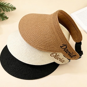 May include: Three straw visors in tan, white, and black are stacked. The tan visor has the name "David" embroidered in black. The white visor has the name "Olivia" embroidered in brown. The visors have a curved brim and an adjustable strap.