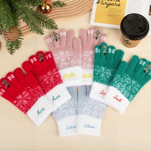 Peut inclure: Une collection de gants d'hiver personnalisés en rouge, rose, vert et bleu clair. Chaque gant présente un motif de flocon de neige, un sapin de Noël sur le bout des doigts et un nom brodé sur le poignet. Les gants sont disposés sur une surface claire.