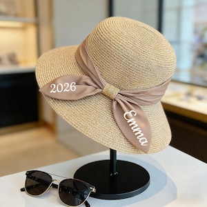 May include: A beige straw sun hat with a wide brim and a brown ribbon. The ribbon is tied in a bow and features the text "2026" and "Emma". The hat is displayed on a black stand, with sunglasses in the foreground.