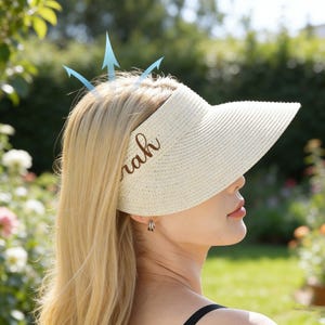 May include: A cream-coloured sun visor with the name "Sarah" in brown script. The visor is made of woven material and has a wide brim. The image shows a person wearing the visor outdoors in a garden setting.
