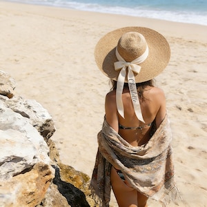 Puede incluir: Un sombrero de paja de ala ancha con una cinta color crema y el nombre "Charlotte" impreso. El sombrero se usa en una playa soleada, combinado con un chal estampado y un bikini. Un accesorio elegante para un día en la playa.