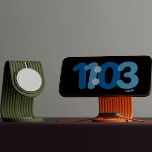 Könnte beinhalten: Ein grüner und weißer Apple Watch-Ladeständer und ein orangefarbener Telefonständer mit einem schwarzen Bildschirm, der die Uhrzeit 11:03 anzeigt. Die Ständer stehen auf einer dunkelbraunen Oberfläche.