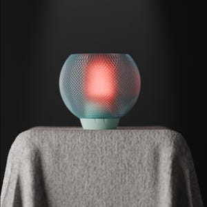 Könnte beinhalten: Eine kugelförmige, türkisfarbene Lampe mit gemusterter Außenseite, die ein warmes, rotes Leuchten ausstrahlt. Sie steht auf einem hellgrünen Sockel, der auf einer grauen, strukturierten Oberfläche platziert ist. Der Hintergrund ist dunkel und einfarbig.