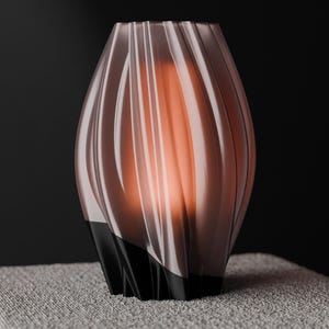Könnte beinhalten: Eine dekorative Vase mit einem einzigartigen, fließenden Design. Die Vase hat eine hellbraune, gerippte Außenseite und einen schwarzen Sockel. Das Innere leuchtet orangefarben und erzeugt eine warme Atmosphäre. Die Vase steht auf einer strukturierten, grauen Oberfläche.