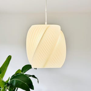 Könnte beinhalten: Cremefarbene Pendelleuchte mit einem einzigartigen, gerippten Design. Die Leuchte hängt an einem weißen Kabel und hat einen weichen, warmen Schein. Die Form der Lampe ist ein abgerundetes, längliches Oval. Eine grüne Pflanze ist unten links zu sehen.