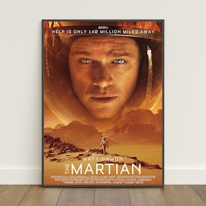 May include: A framed movie poster for The Martian featuring a large close-up of a person in a space helmet against an orange landscape. The text reads HELP IS ONLY 140 MILLION MILES AWAY, MATT DAMON, and THE MARTIAN, with small credits at the bottom.