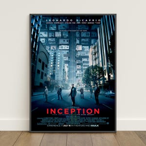 Puede incluir: Un póster de película enmarcado de Inception que muestra un paisaje urbano azul oscuro donde los edificios se pliegan hacia arriba. El título Inception está en letras rojas negritas abajo, con nombres del reparto y fecha en blanco.