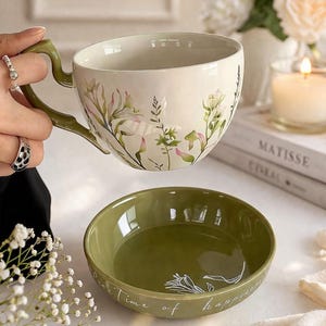 Peut inclure: Un ensemble tasse à thé et bol vert olive. La tasse à thé présente un motif floral avec des accents verts et roses. Le bol porte l'inscription "Pure Time of happiness" et un motif floral. La tasse à thé est tenue au-dessus du bol.
