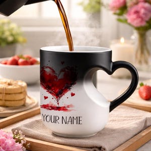 May include: A white mug with a black handle and rim is being filled with a dark liquid. The mug features a red heart design and the text "YOUR NAME". The handle is heart-shaped.