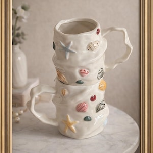 Peut inclure: Deux tasses empilées en céramique blanche, décorées de coquillages et d'étoiles de mer. Les tasses ont des anses ondulées et des accents colorés. Les tasses sont exposées sur une table en marbre.