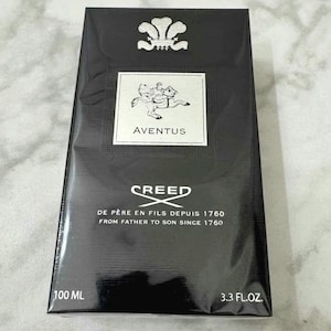 Puede incluir: Caja rectangular negra de colonia Creed Aventus. La caja presenta el logotipo de Creed, un escudo y el texto "AVENTUS". También incluye el texto "DE PÈRE EN FILS DEPUIS 1760". Contiene 100 ml / 3.3 FL.OZ.
