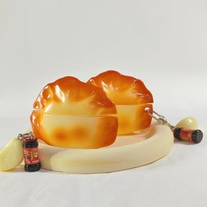 May include: Two orange and yellow dumpling-shaped containers sit on a cream-colored, oval-shaped plate. Small, detailed keychains of a garlic clove and a bottle of sauce are attached to the plate.