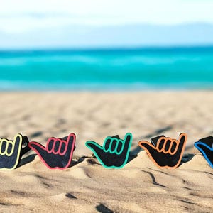 May include: Five shaka hand sign ornaments arranged in a row, each featuring a black center with a distinct colored outline in yellow, red, green, orange, and blue. These decorative pieces display the iconic gesture with extended thumb and pinky.